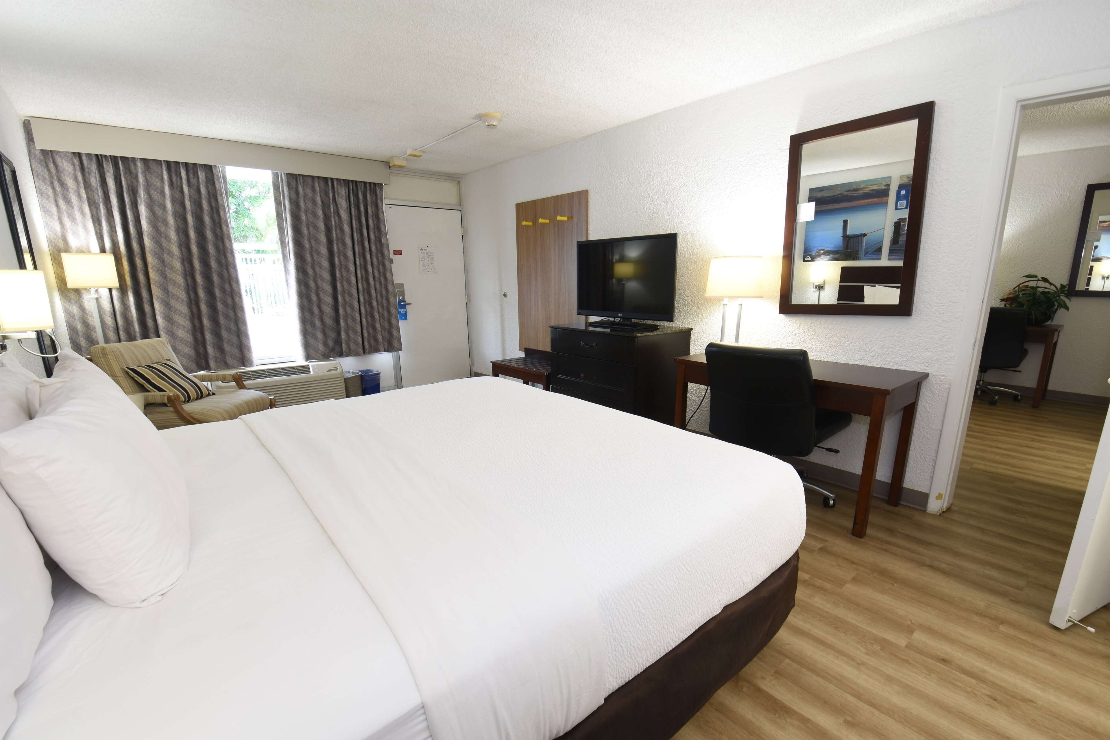 A hotel room with a large bed, a desk with a television, a chair, and a desk lamp.