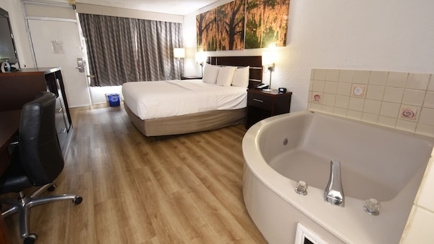 A hotel room with a bed, a desk, a chair, a desk lamp, a nightstand, a telephone, a clock, a painting, and a bathtub.