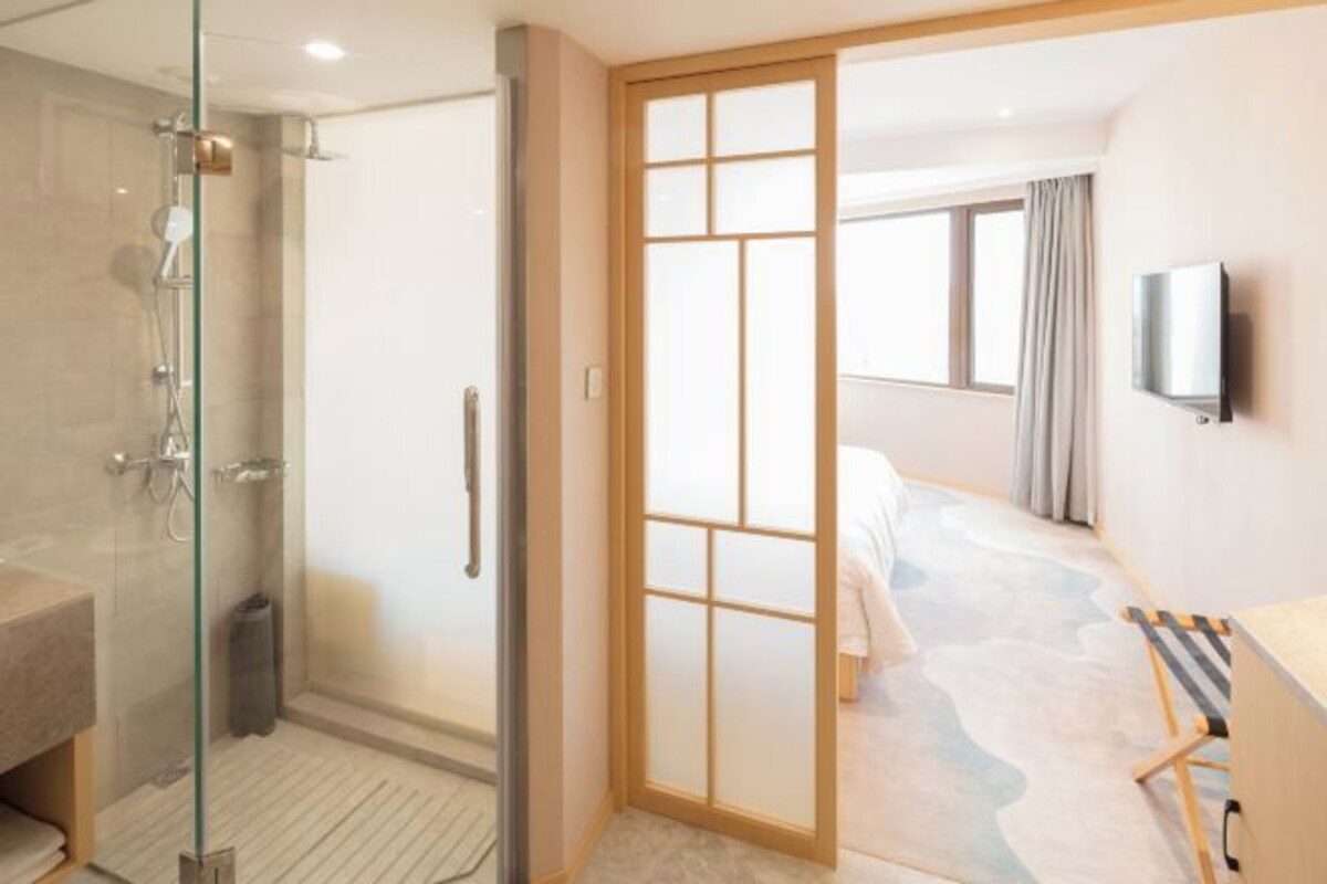 Habitación de hotel con cabina de ducha de vidrio, puerta corredera de madera, cama con cabecera de color claro, televisor de pantalla plana montado en la pared y ventana con cortinas.