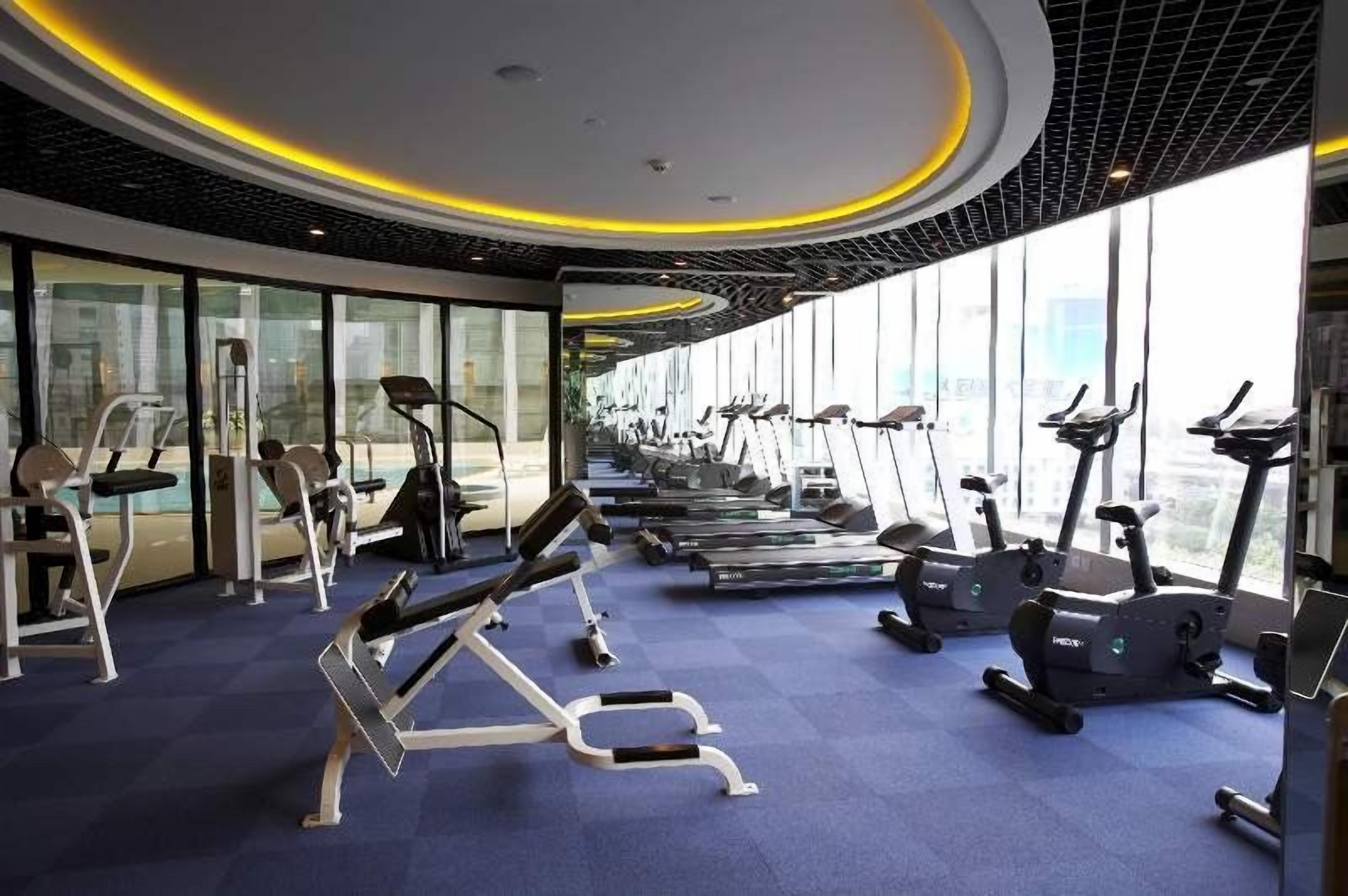 A modern gym with cardio machines, weight benches, and a large window offering a city view.