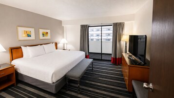 Habitación de hotel con cama, mesitas de noche, escritorio con televisor, banco y vista al exterior.