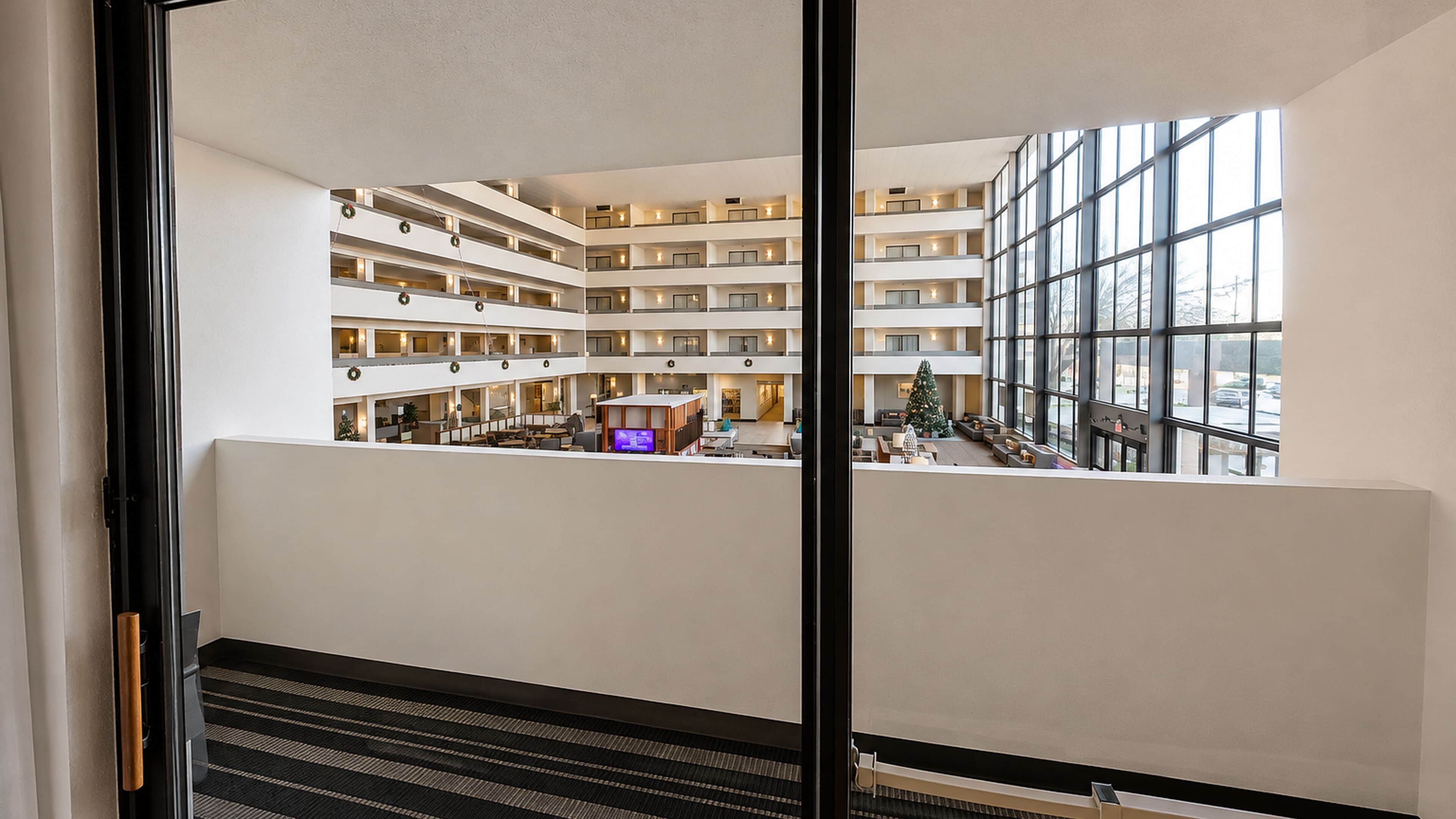 Un vestíbulo de hotel de varios niveles con un área de estar central, un árbol de Navidad y vista al estacionamiento a través de grandes ventanales.
