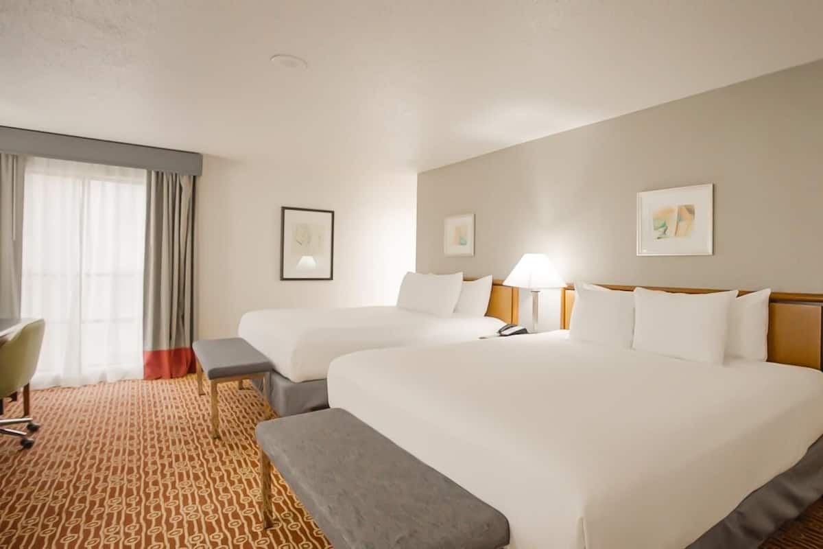 A hotel room with two beds, a desk, a chair, a lamp, and artwork on the walls.