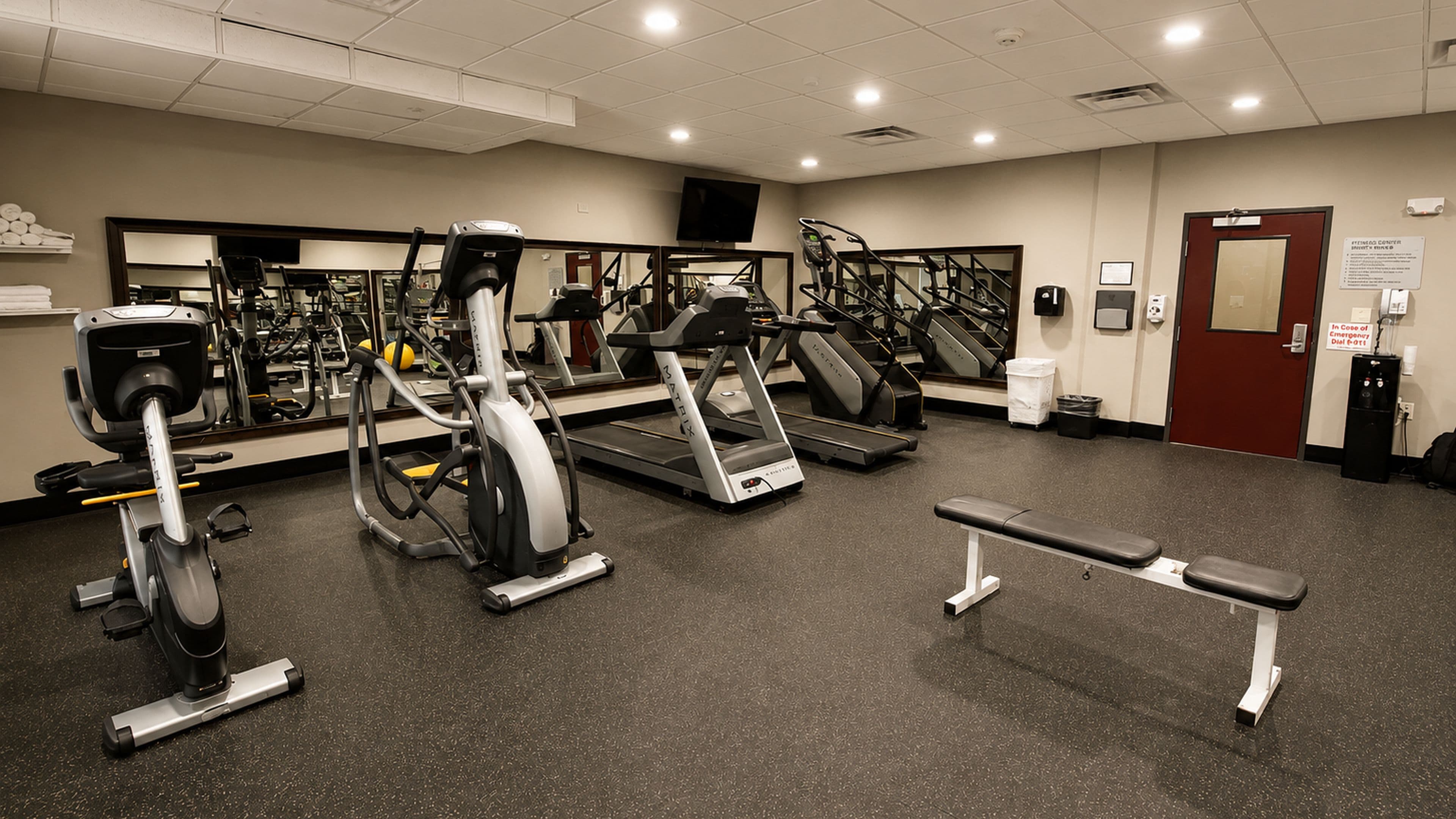 A gym with various exercise equipment, a bench, and a door with an emergency exit sign.