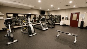 Un gimnasio con diversos equipos de ejercicio, una banca y una puerta con letrero de salida de emergencia.