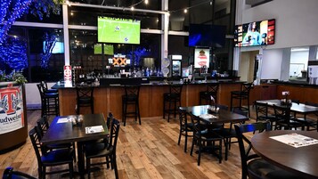 Uma área de bar com diversas opções de assento, um balcão com vários tipos de bebidas e uma televisão exibindo uma partida esportiva.
