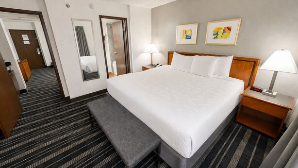 Une chambre d’hôtel avec un grand lit, des tables de chevet, un banc, un miroir et deux œuvres d’art encadrées accrochées au mur.