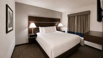 A hotel room with a large bed, two bedside tables with lamps, a headboard, a window with curtains, and a flat-screen TV.