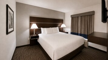 A hotel room with a large bed, two bedside tables with lamps, a headboard, a window with curtains, and a flat-screen TV.