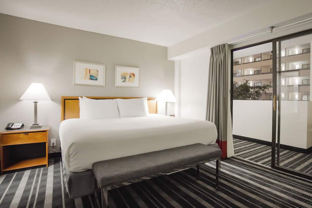 A hotel room with a bed, bedside lamps, a bench, a view of the building, and a window with curtains.
