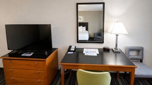 Une chambre d’hôtel avec un téléviseur posé sur une commode en bois, un bureau avec une lampe, une chaise et un miroir au mur.