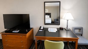 Habitación de hotel con televisor sobre una cómoda de madera, un escritorio con lámpara, una silla y un espejo en la pared.