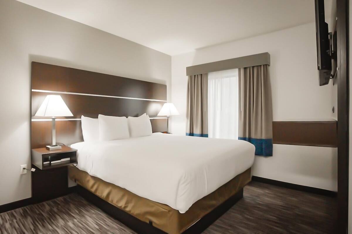 A hotel room with a large bed, bedside lamps, a nightstand, a window with curtains, and a flat-screen TV mounted on the wall.