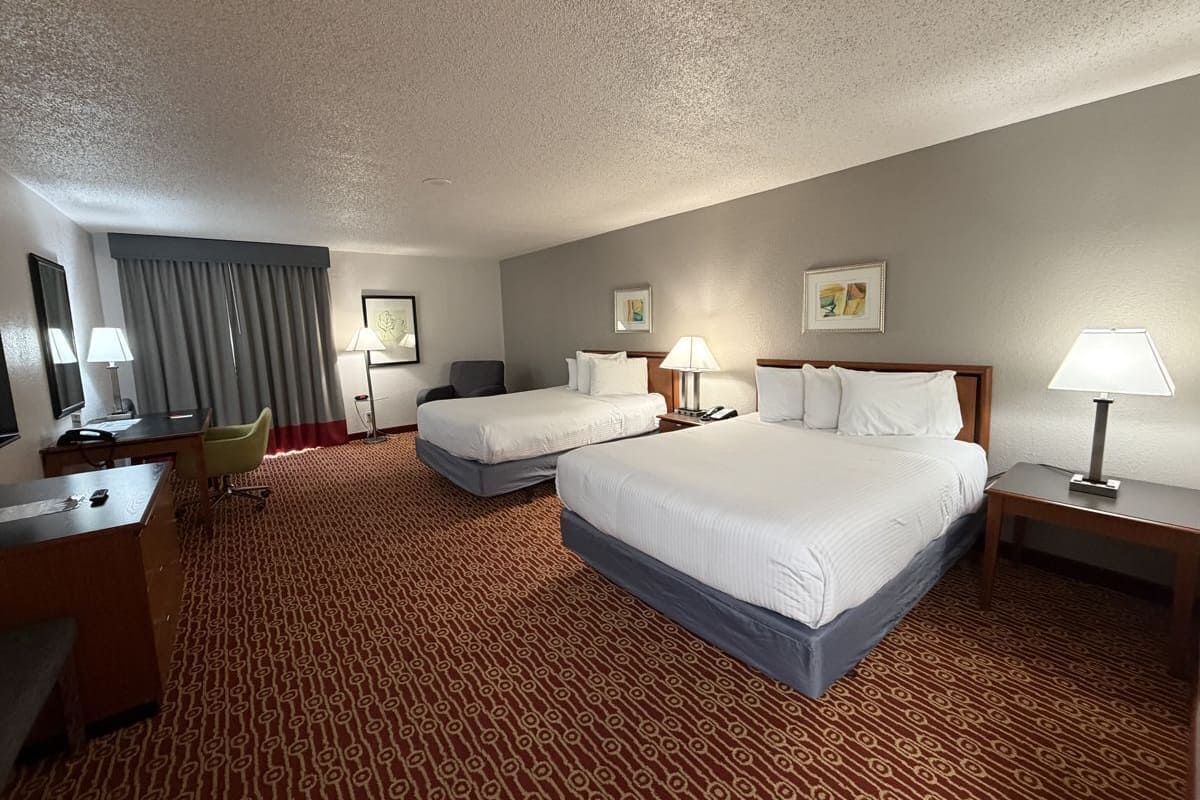 A hotel room with two beds, a desk, a chair, and a lamp.