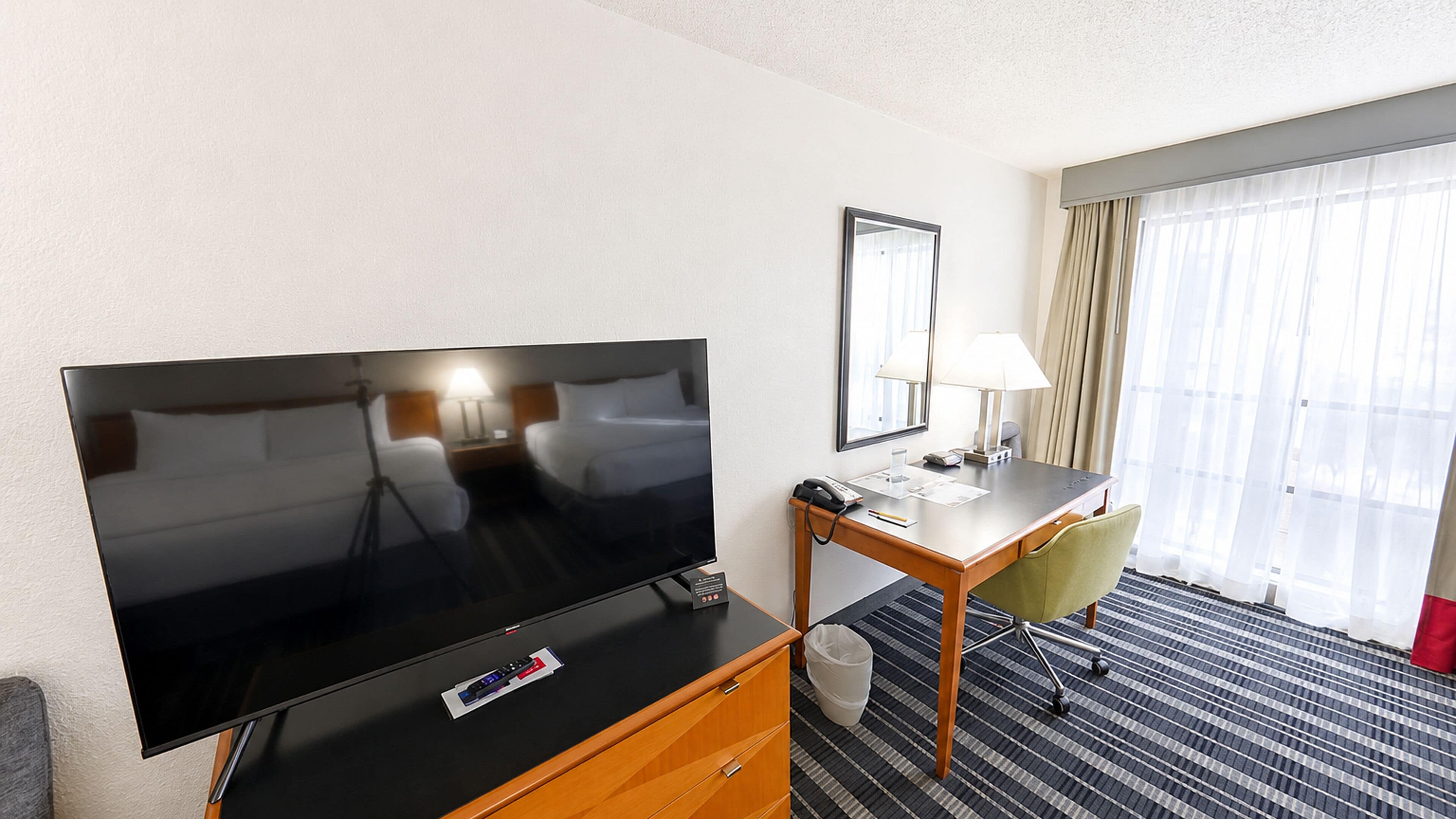 Quarto de hotel com televisão de tela plana, escrivaninha com luminária, cadeira e uma janela grande com cortinas.