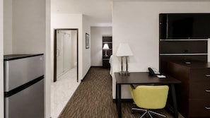 Habitación de hotel con refrigerador, escritorio con lámpara, una silla, televisor y cama al fondo.