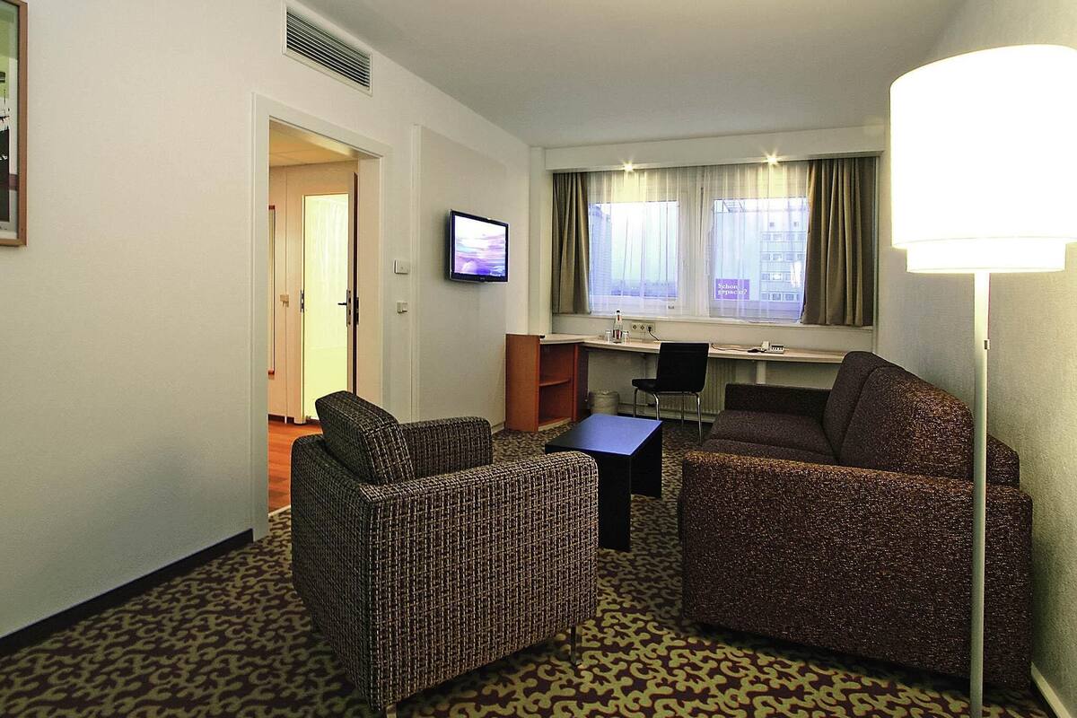 Quarto de hotel com sofá, poltrona, escrivaninha e televisão.
