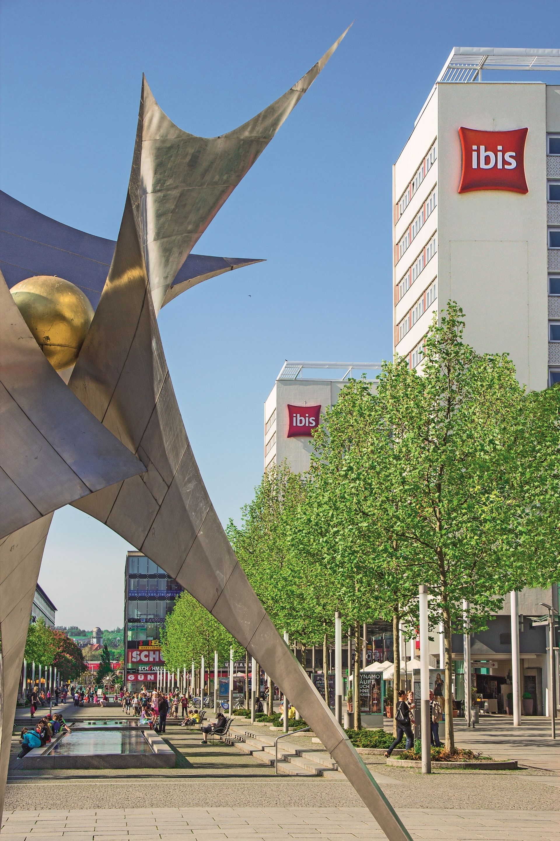 A modern urban plaza with a prominent metallic sculpture, a large 'ibis' hotel sign, and a clear blue sky.