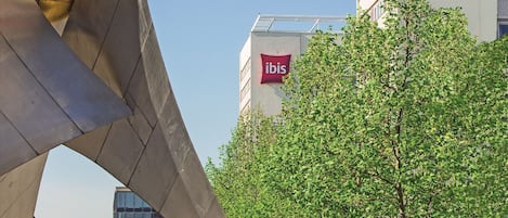 A modern urban plaza with a prominent metallic sculpture, a large 'ibis' hotel sign, and a clear blue sky.
