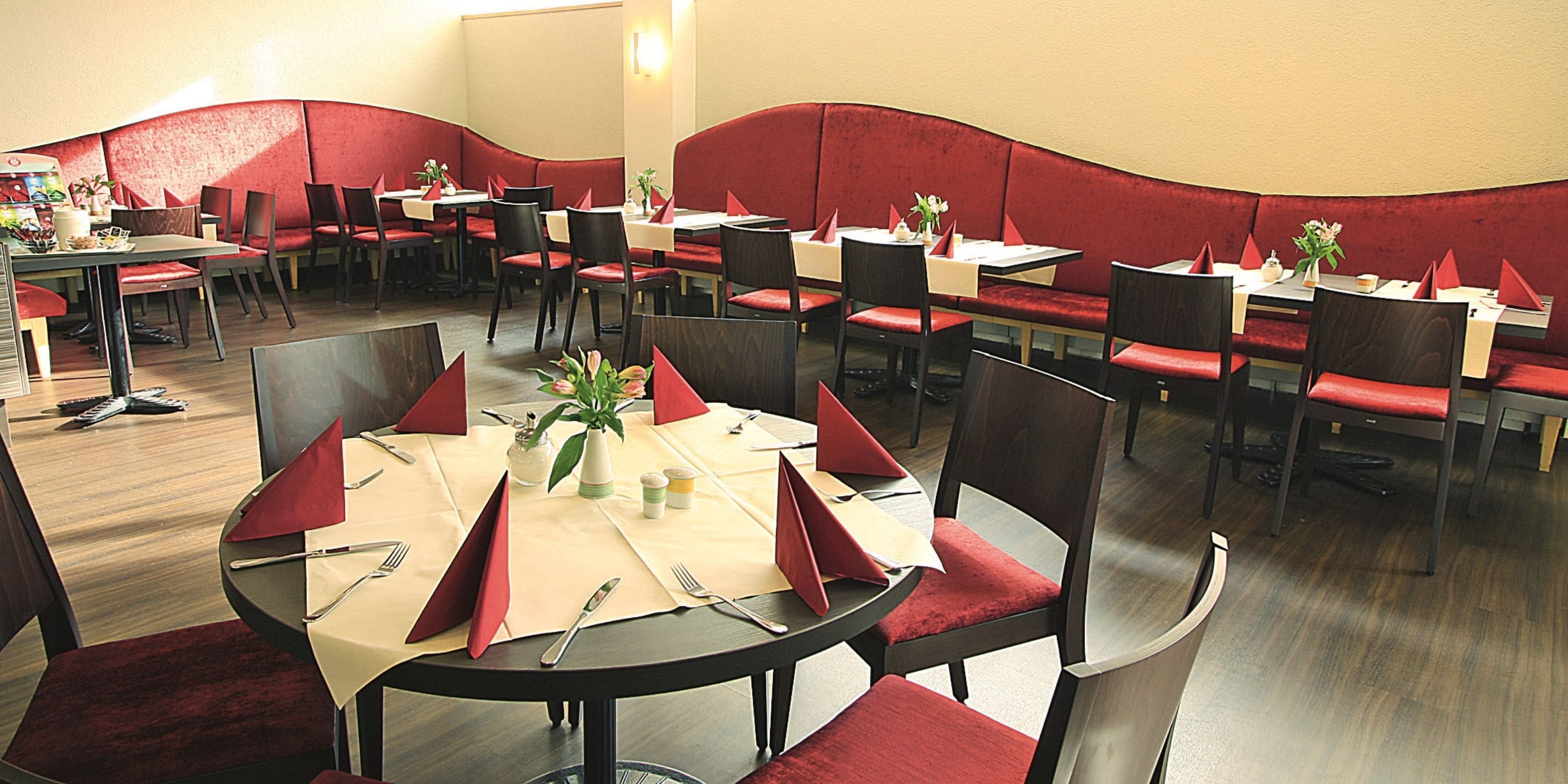A restaurant with red cushioned booths and tables, each set with cutlery and napkins.