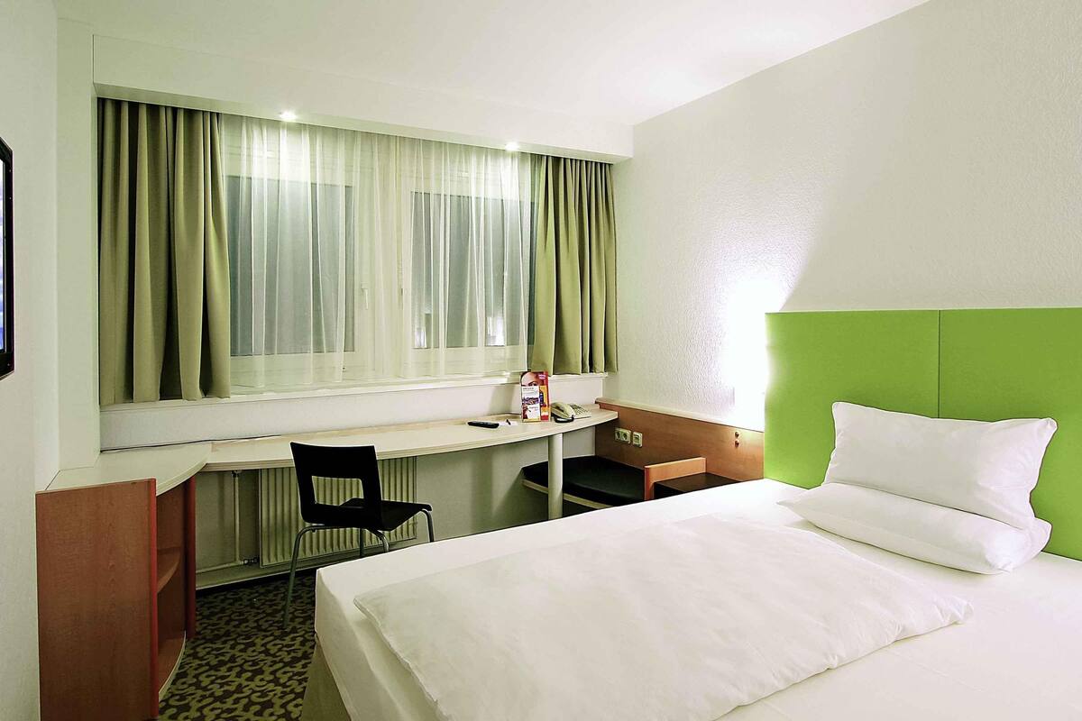 Quarto de hotel com cama, escrivaninha, cadeira, telefone e janela com cortinas.