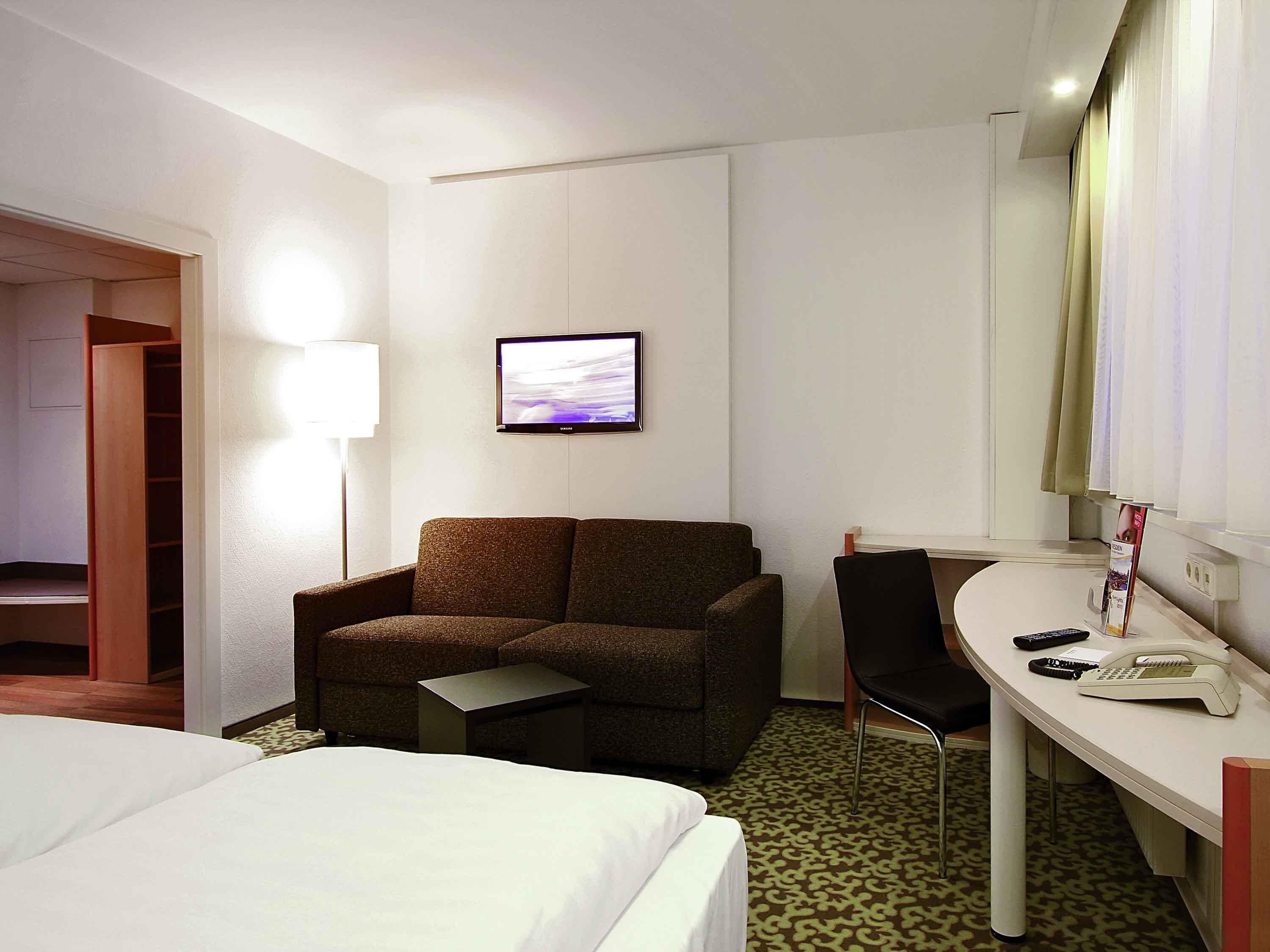 A hotel room with a bed, a sofa, a desk with a telephone, a chair, a television mounted on the wall, and a patterned carpet.