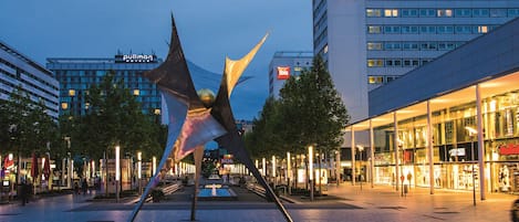 A modern hotel with a prominent 'ibis' sign, a large sculpture in the foreground, and a well-lit urban plaza with trees and shops.