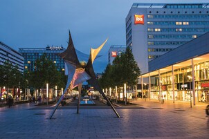 Un hotel moderno con un llamativo letrero de 'ibis', una gran escultura en primer plano y una plaza urbana bien iluminada con árboles y tiendas.