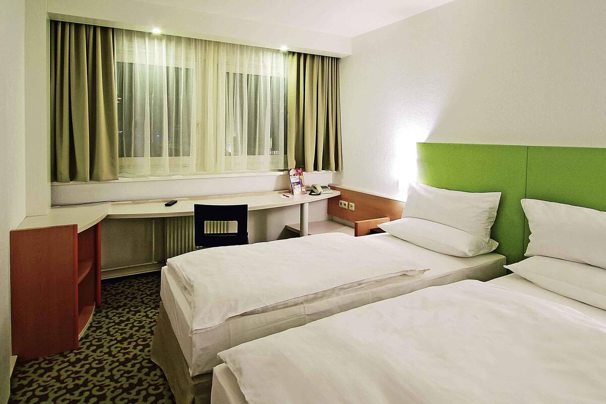 Quarto de hotel com duas camas, uma escrivaninha, uma cadeira, janela com cortinas e cabeceira verde.
