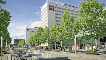 Ein moderner Stadtplatz mit einem Brunnen, Bäumen und Gebäuden, darunter ein ibis Hotel.