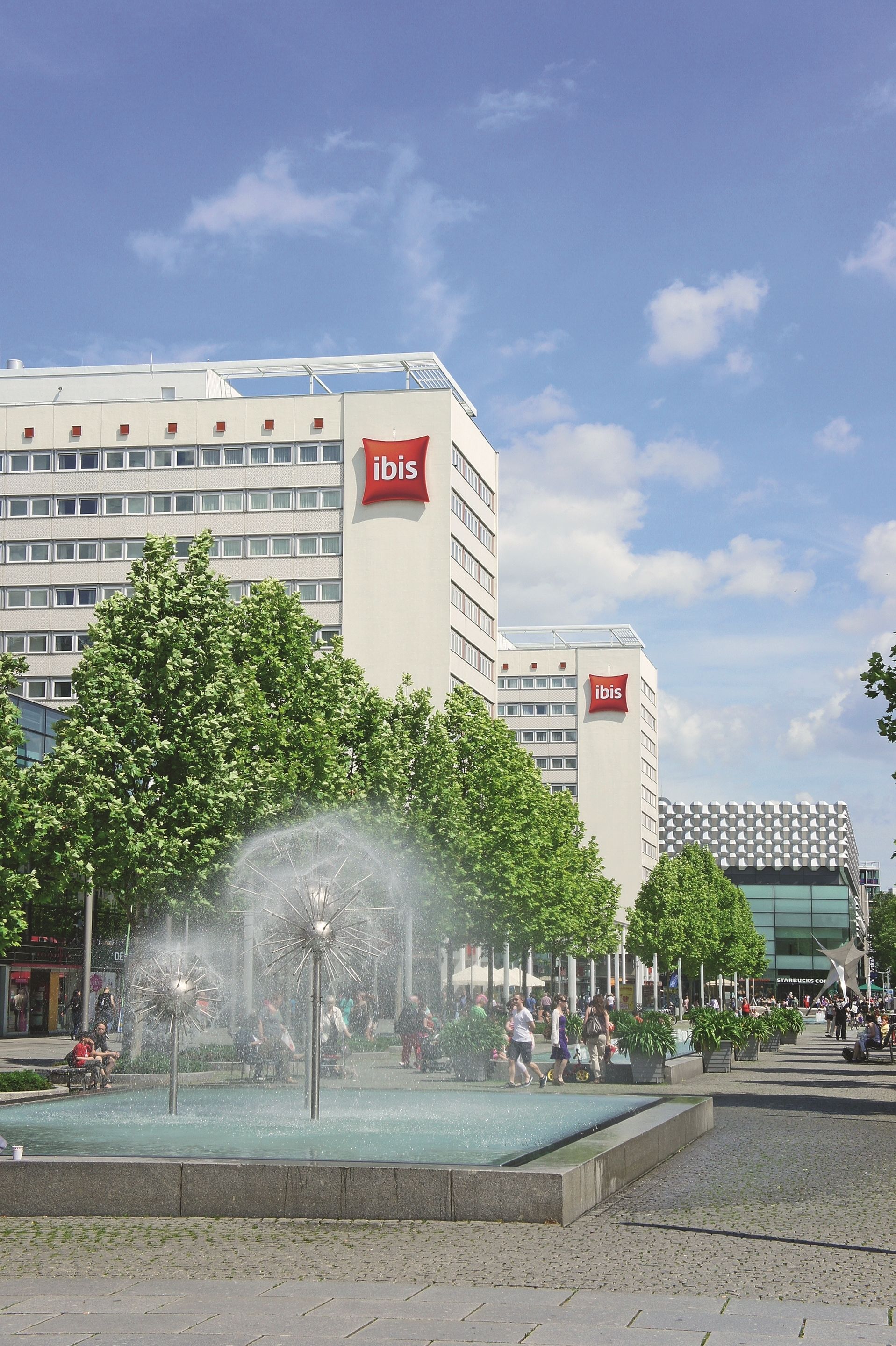 A modern hotel with a prominent ibis logo, a central fountain, and a Starbucks nearby.