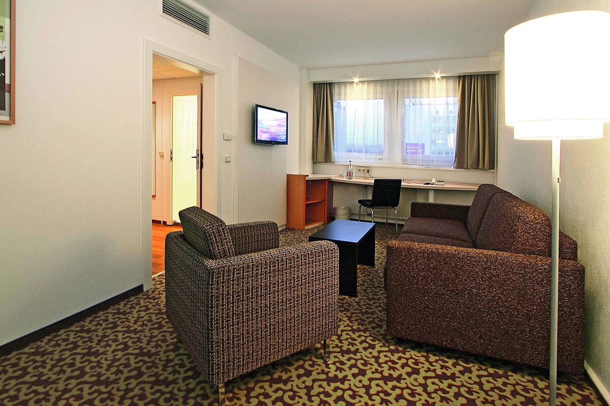 Habitación de hotel con sofá, dos sillones, escritorio, televisor y ventana con cortinas.