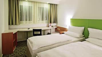 A hotel room with two beds, a desk, a chair, a window with curtains, and a green headboard.