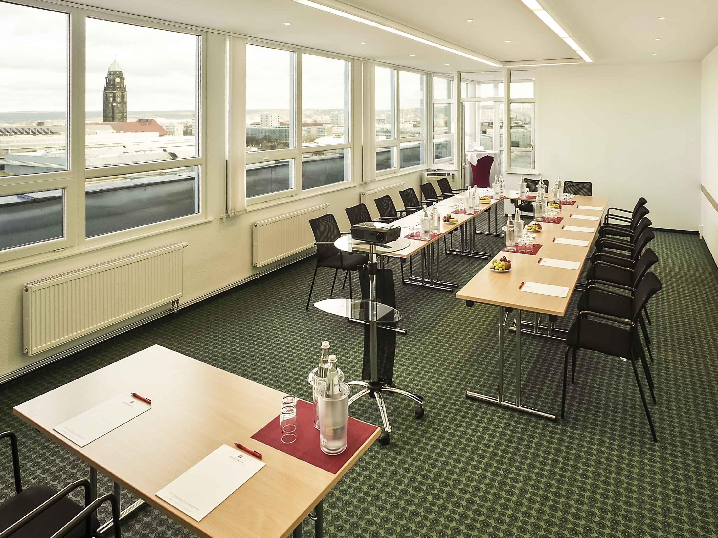 Ein Konferenzraum mit großen Fenstern, einem langen Tisch, Stühlen und Blick auf eine Stadtlandschaft.
