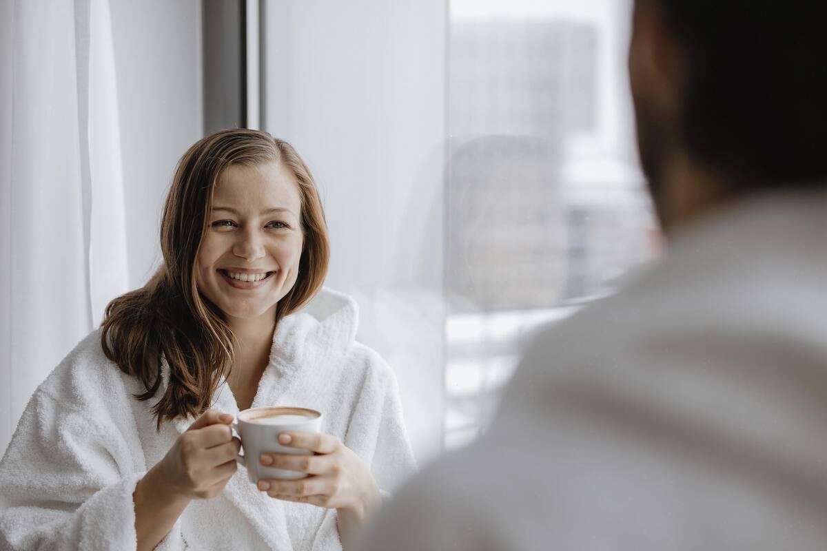 Una persona con bata blanca sostiene una taza, sentada junto a una ventana con vistas a edificios.
