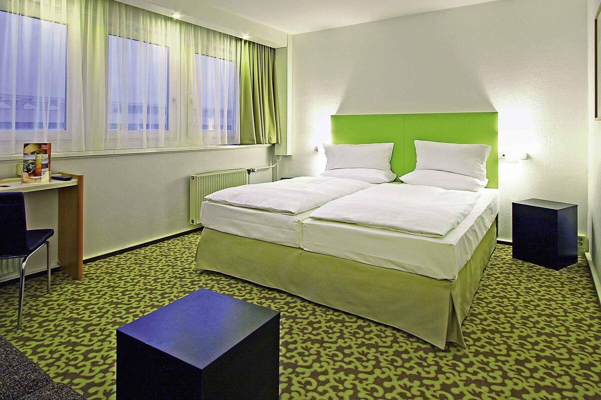 Quarto de hotel com uma cama de casal, escrivaninha, cadeira, mesinha e radiador.