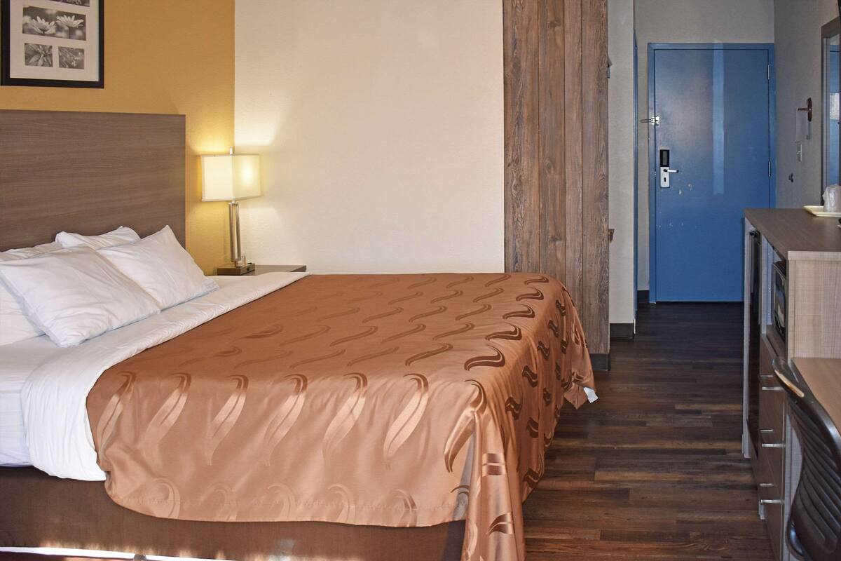 Une chambre d’hôtel avec un lit, une table de chevet avec une lampe, une tête de lit en bois, une porte bleue et un coin cuisine.