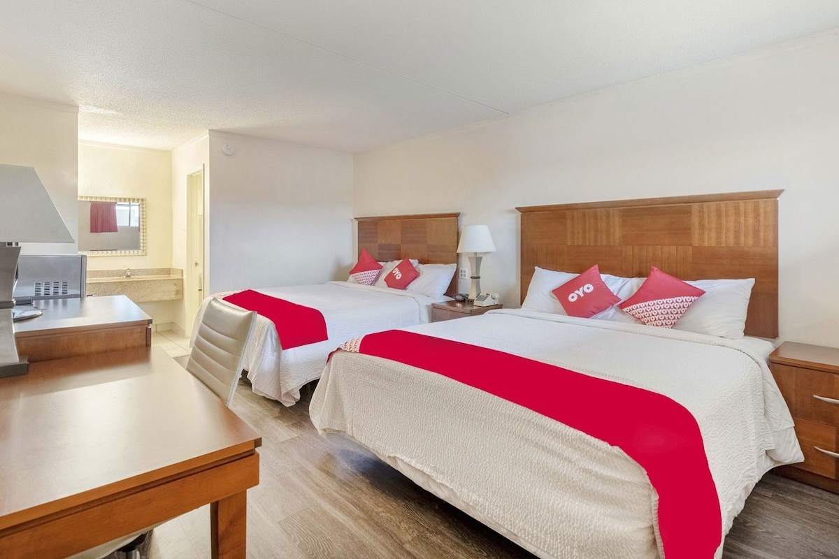 Quarto de hotel com duas camas, uma mesa, uma cadeira, um abajur e um banheiro visível ao fundo.