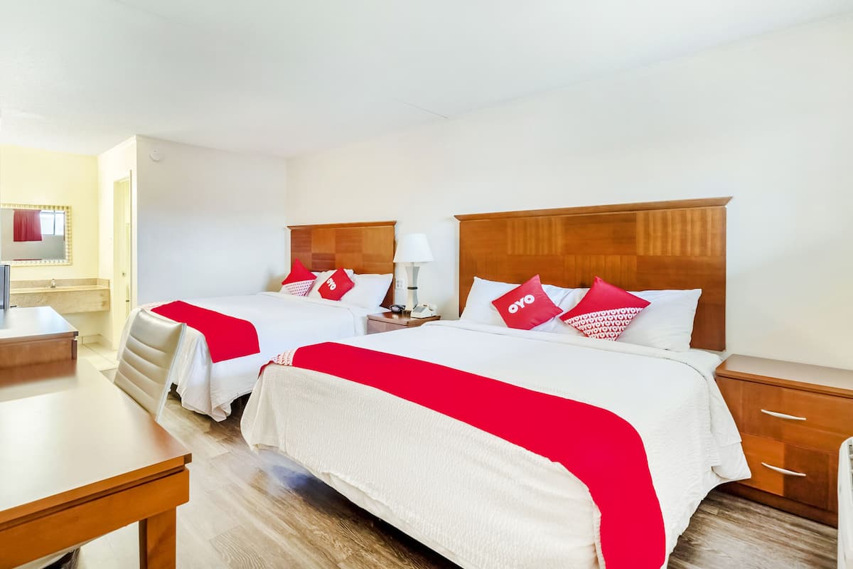 A hotel room with two beds, each with a wooden headboard, white bedding, and red pillows. There is a desk with a chair, a nightstand with a lamp, and a wooden dresser.