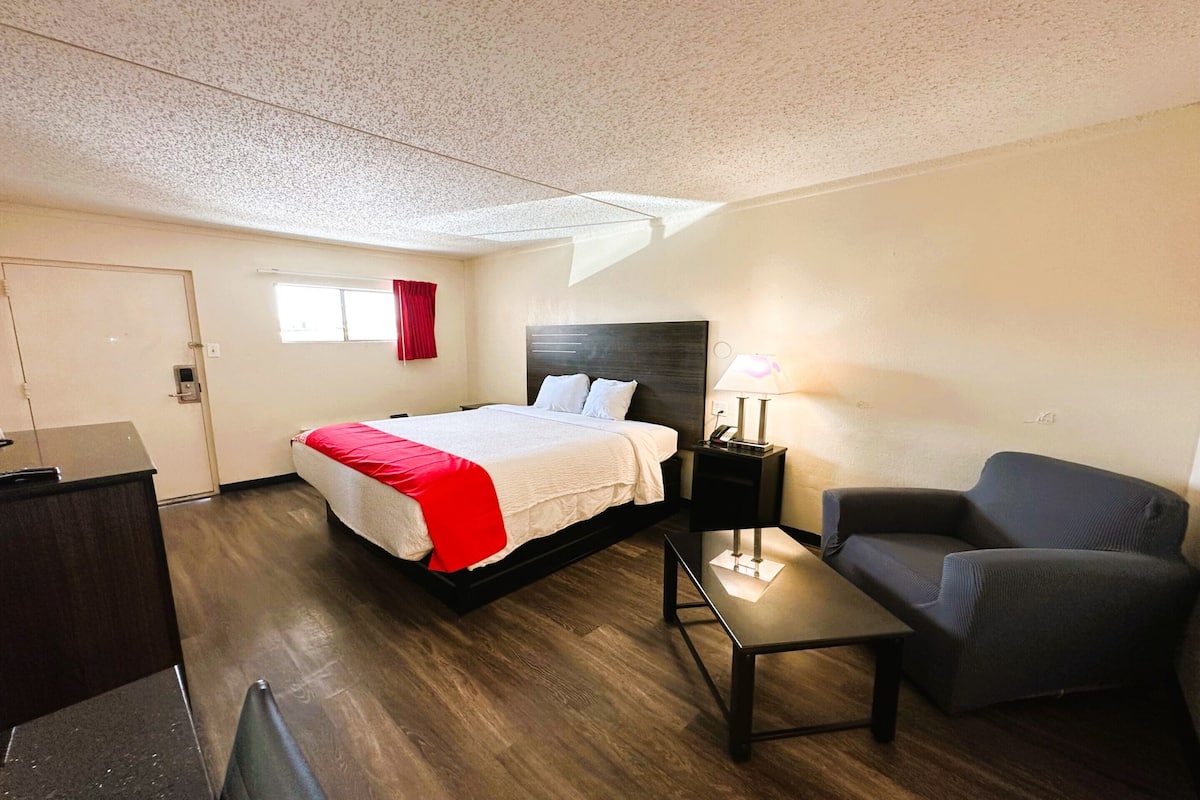 A hotel room with a bed, a desk, a chair, and a nightstand with a lamp.