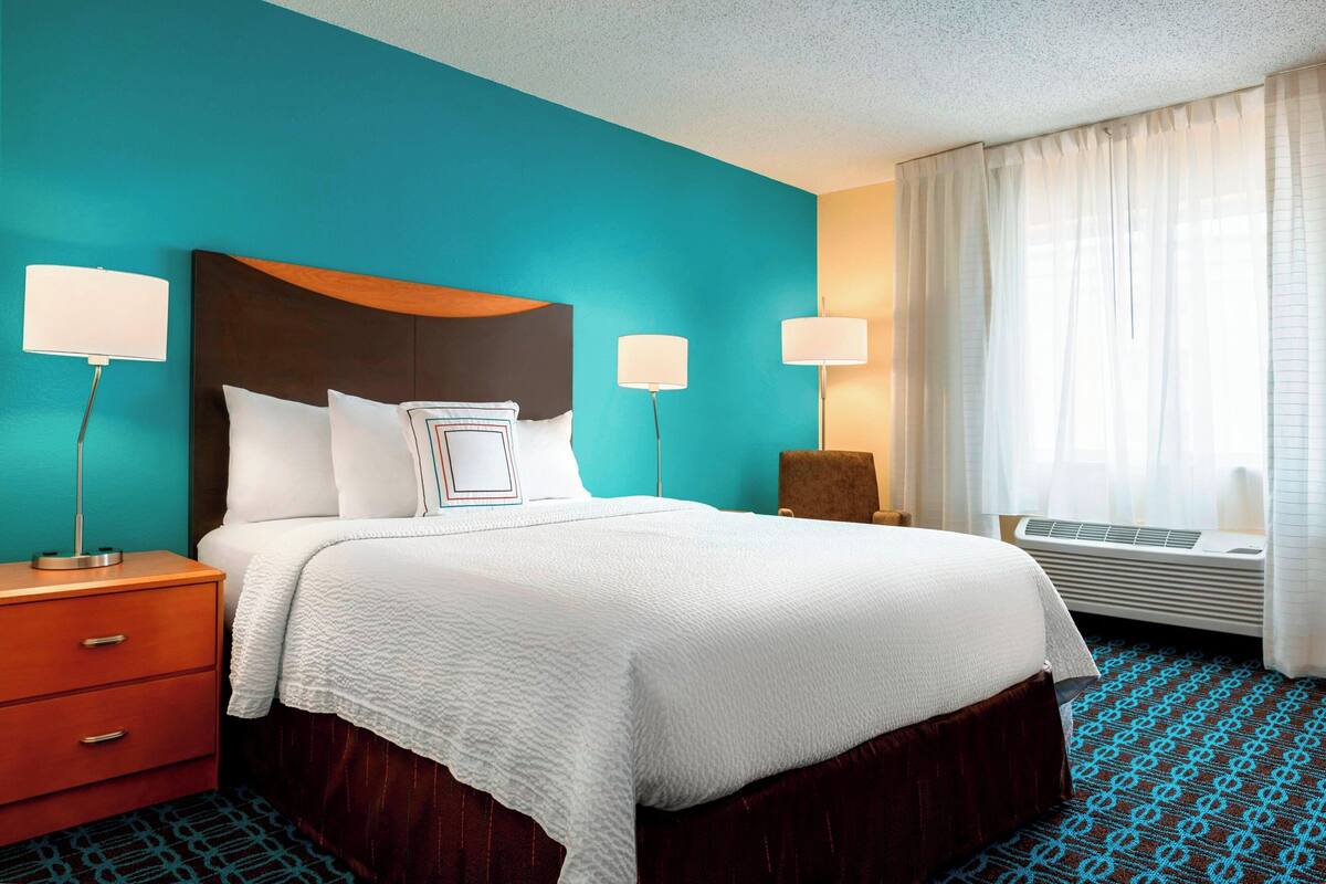 A hotel room with a bed, a nightstand, two bedside lamps, a chair, and a window with curtains.