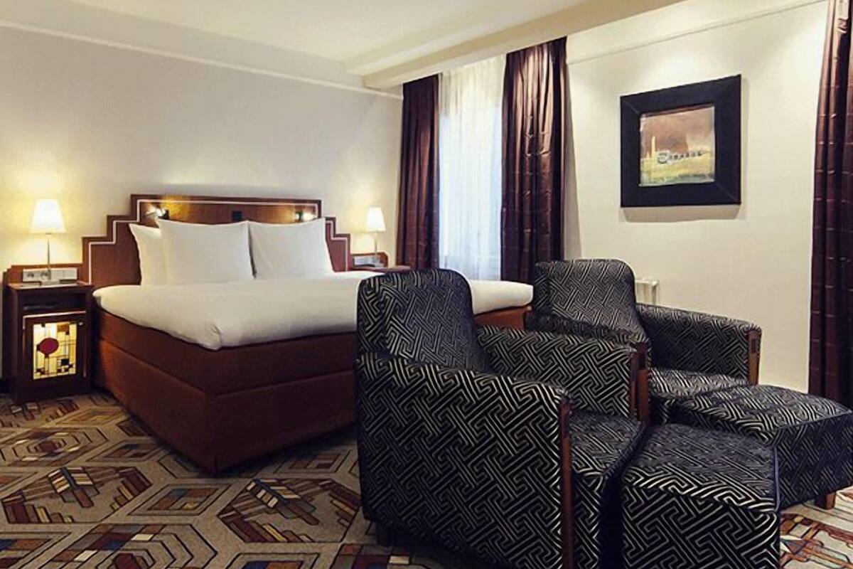 A hotel room with a bed, two armchairs, a nightstand, a lamp, a framed picture on the wall, and a patterned carpet.