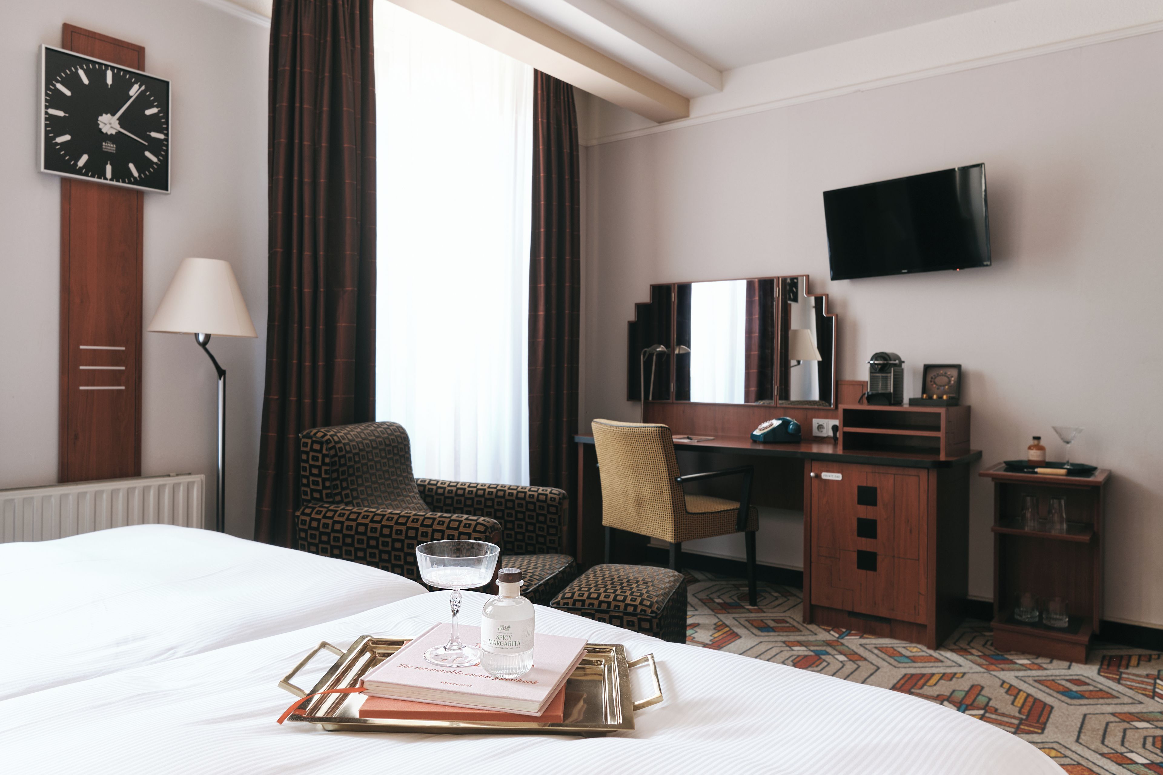 A hotel room with a bed, a desk, a chair, a television, and a clock on the wall.