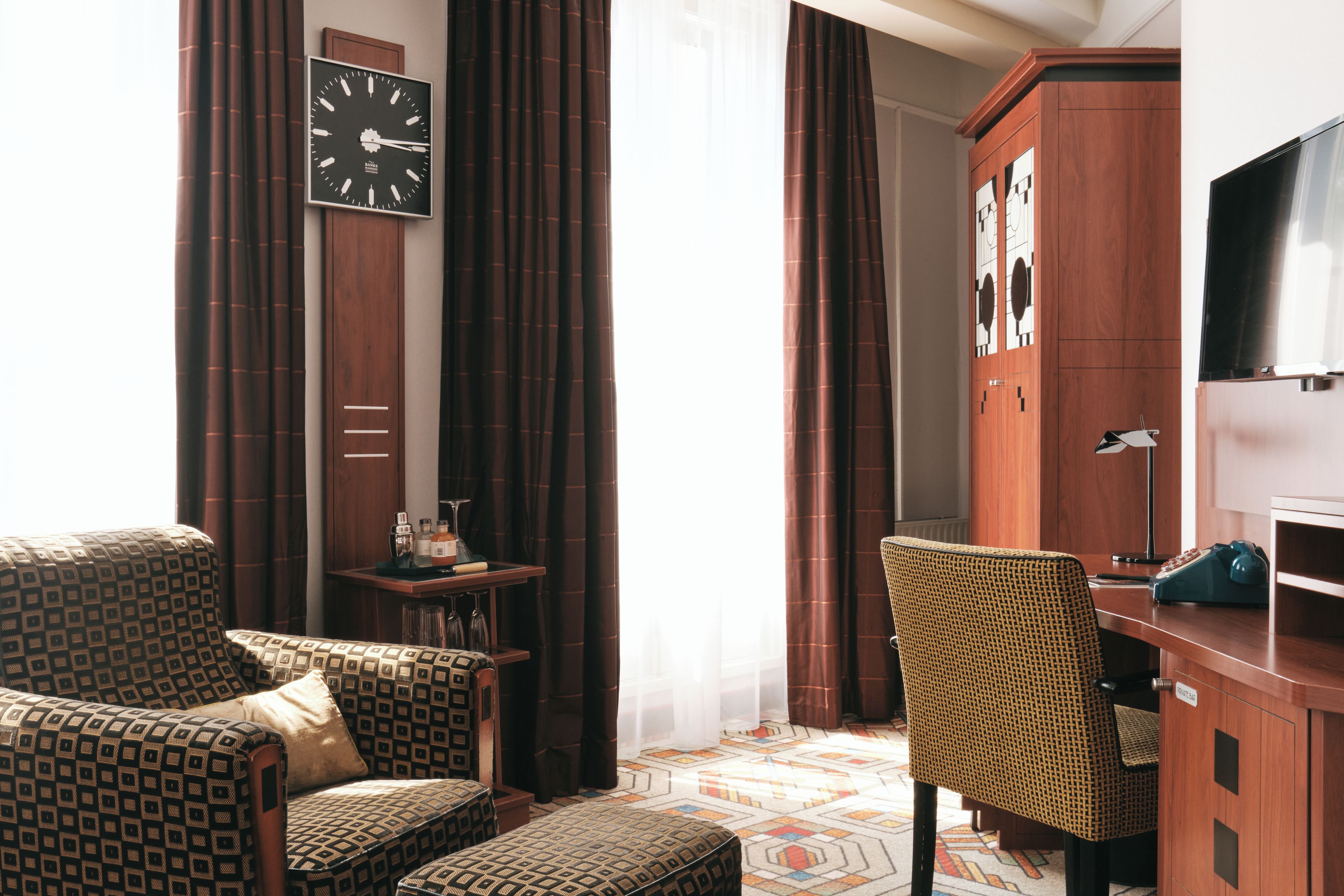 A hotel room with a bed, two patterned armchairs, a small table with a lamp, a television, and a clock on the wall.