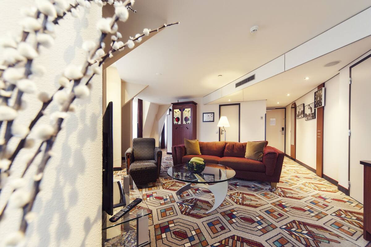 A hotel room with a patterned carpet, a sofa, a chair, a glass coffee table, and a television.