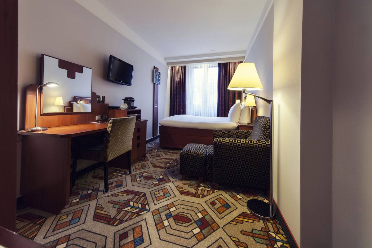 A hotel room with a bed, desk, chair, television, and a patterned carpet.