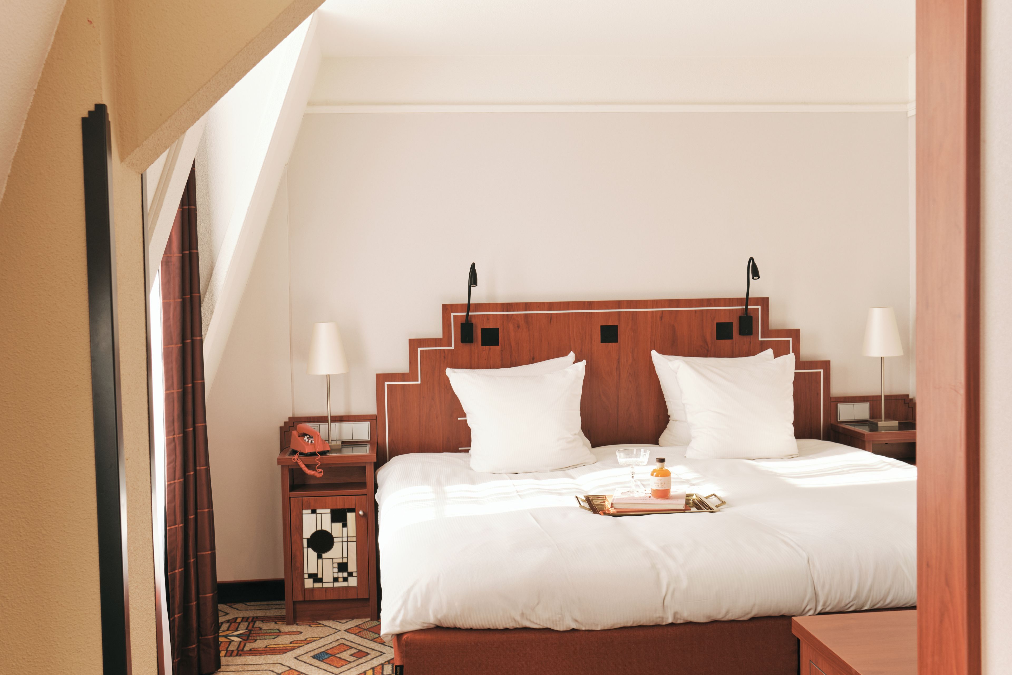 A hotel room with a wooden headboard, white bedding, a bedside table with a lamp, and a small decorative item on the bed.
