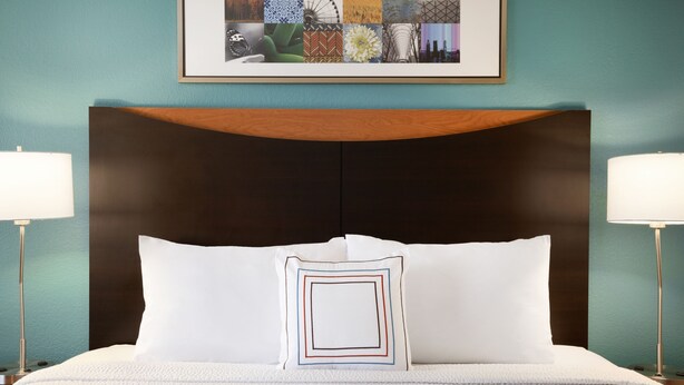 Un lit bien fait, avec une literie blanche et un coussin décoratif, une tête de lit en bois et une œuvre d’art encadrée au-dessus.