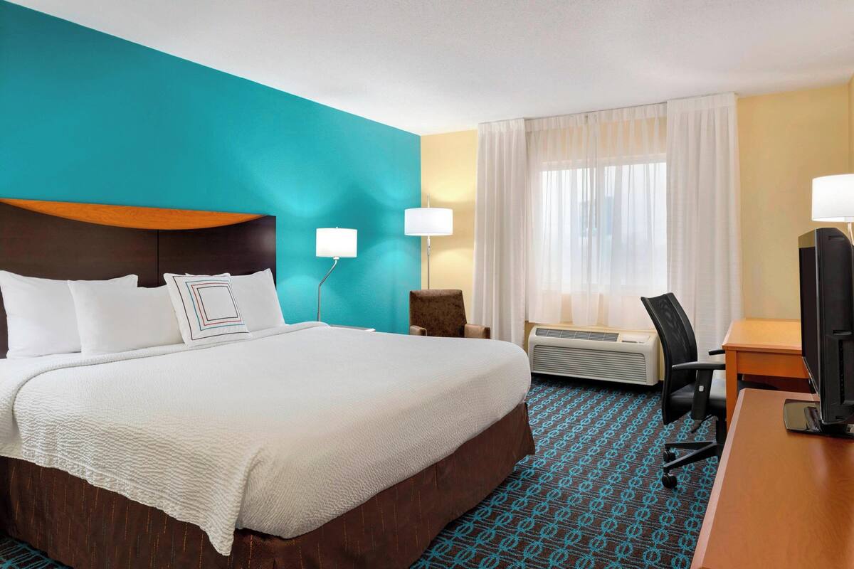 Een hotelkamer met een groot bed, een bureau met een computer, een stoel, een lamp en een airconditioning.