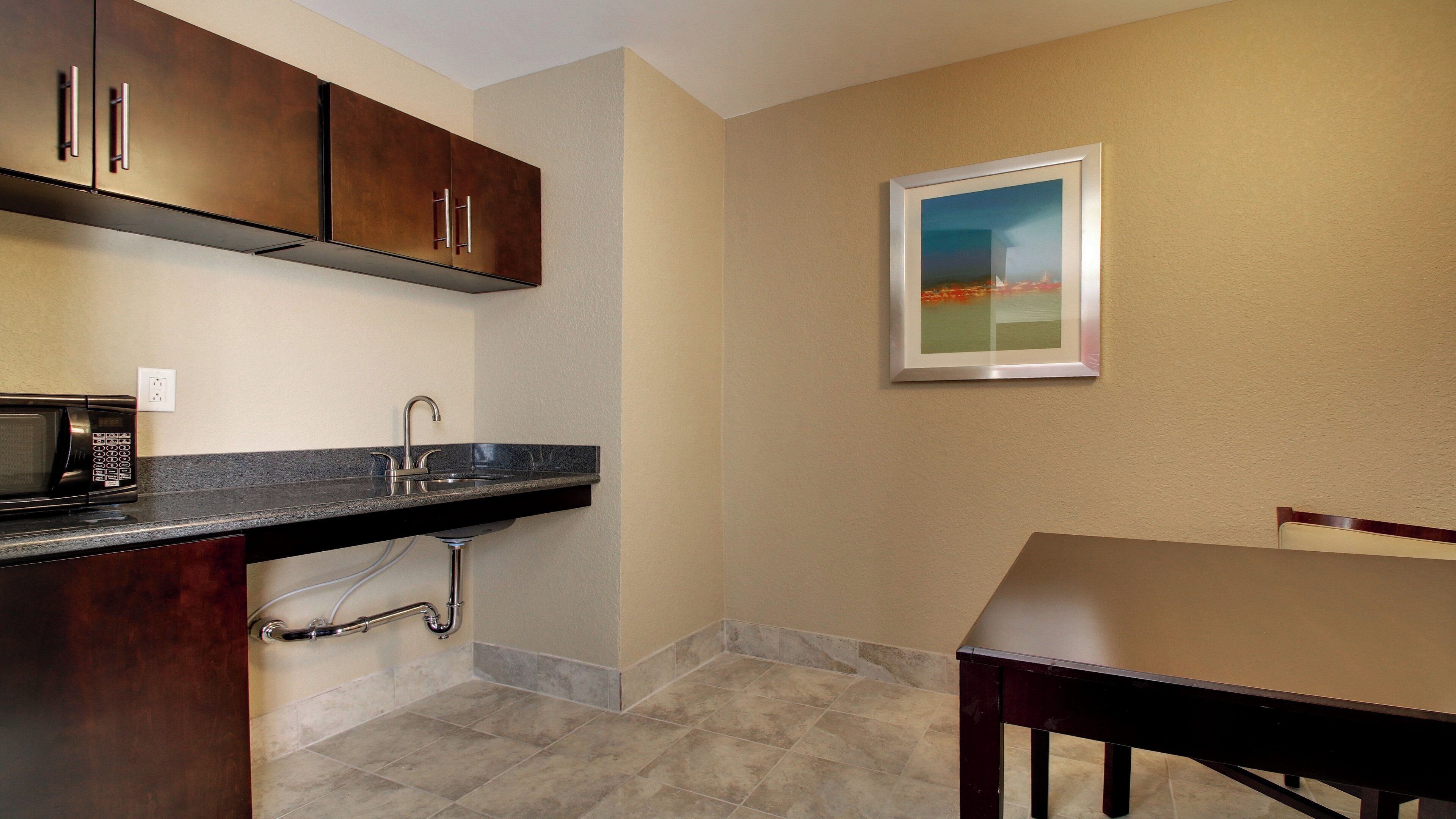A kitchen with a microwave, sink, and cabinets. There is a painting on the wall and a table with chairs.